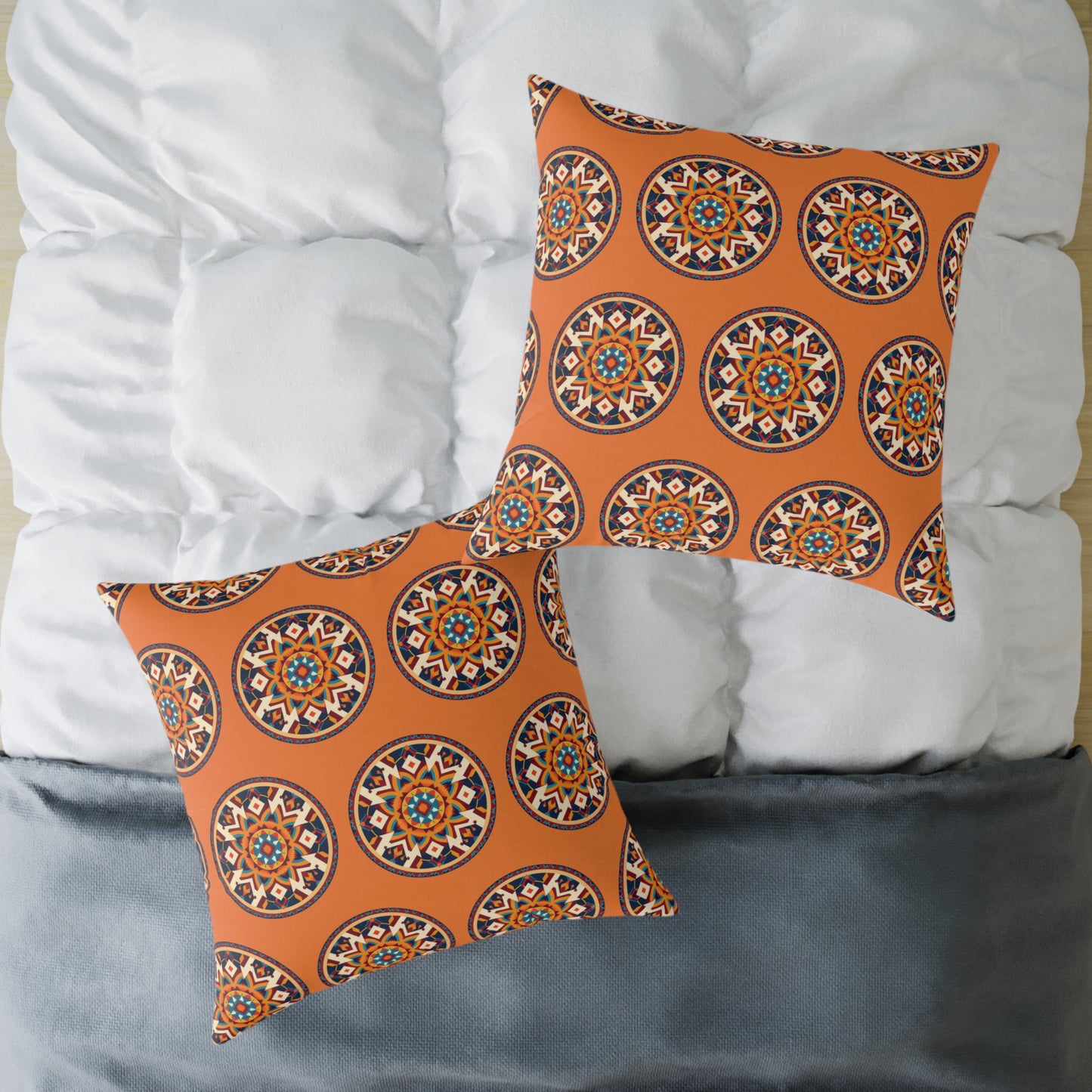 Two square, African-inspired pillows with orange covers featuring a circular, intricate mandala pattern in blue, white, and black rest on a neatly made bed with a white comforter and gray blanket, adding colorful accents to the home decor.