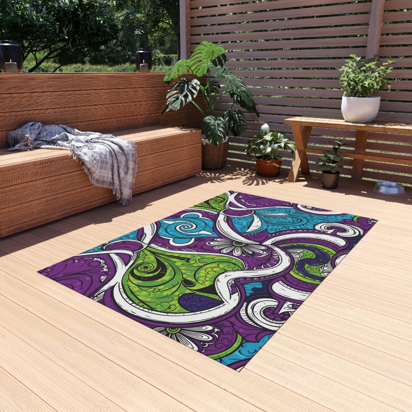 A patio with wooden decking features a wicker sofa draped with a gray throw, a bench with potted plants, and a large, weather-resistant rug in purple, green, and blue swirls. Lush green plants and sunlight complete the cozy outdoor setting.