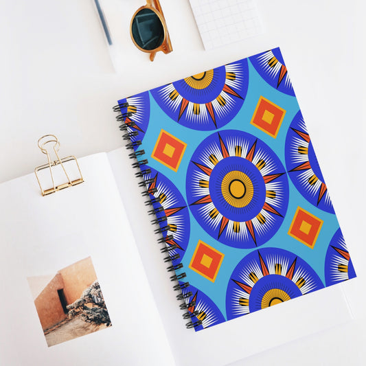 A vibrant African-inspired spiral notebook with blue, orange, and yellow geometric patterns lies on a white desk. Paired with an open book, gold binder clip, sunglasses, and a grid notepad, it offers a premium writing experience in a bright, minimalist scene.