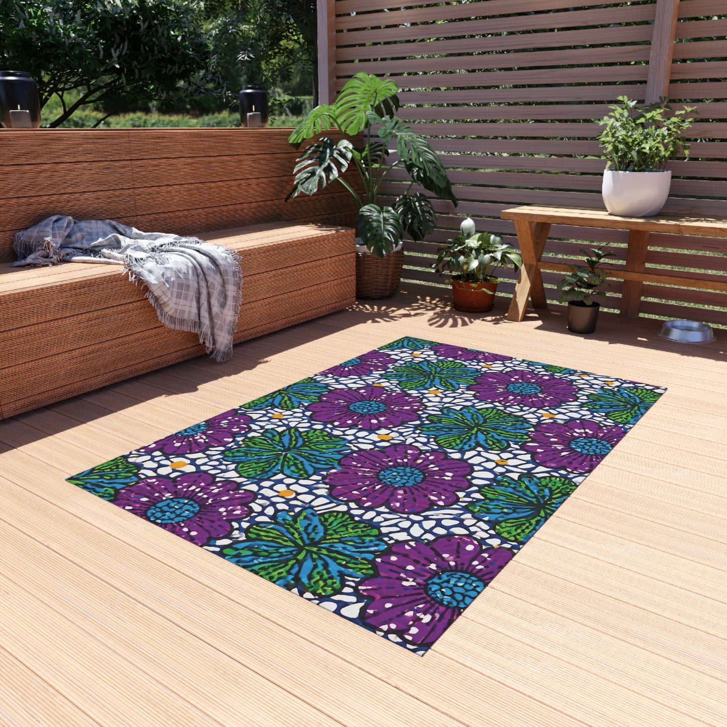A cozy outdoor deck with wooden flooring features a wicker bench, potted plants, and a small table. A vibrant weather-resistant rug with purple, green, and blue floral patterns adds color as sunlight filters through a slatted wood privacy screen.