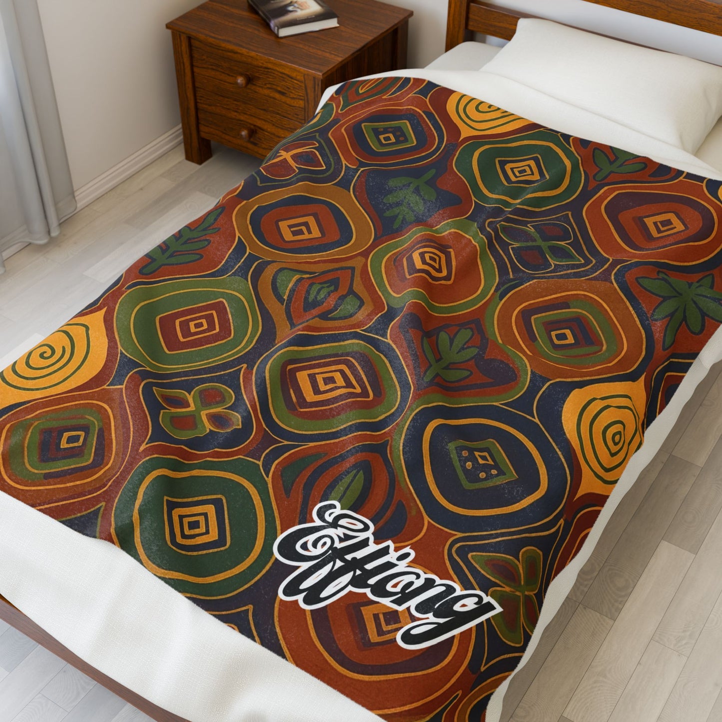 A single bed with an African-inspired blanket featuring abstract, concentric geometric shapes in orange, green, brown, and yellow hues. The blanket also has a white script that says Efflang at the foot. The bed is next to a wooden nightstand and a closed book.