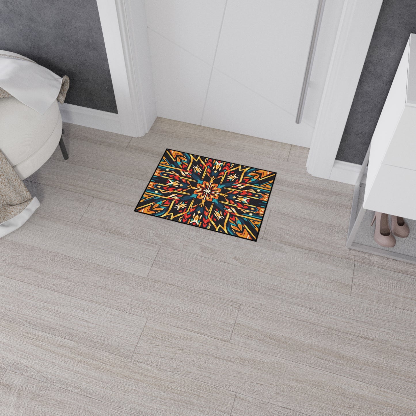 A small rectangular custom floor mat with an African-inspired geometric and floral pattern in red, yellow, blue, and black sits on a light wooden floor near a white door. The mat features non-slip rubber backing for added safety.