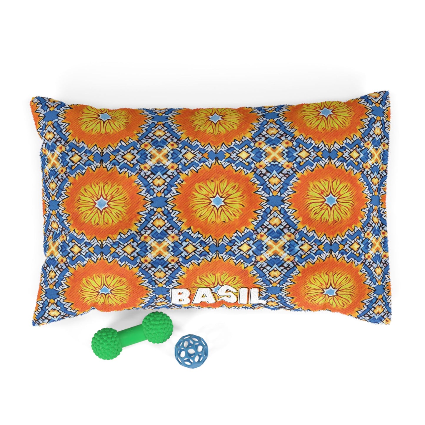 A custom pet pillow features a vibrant blue, orange, and yellow geometric floral pattern with BASIL in bold white letters. In front of this stylish dog bed are two toys: a green bone-shaped chew toy and a blue lattice ball. White background.