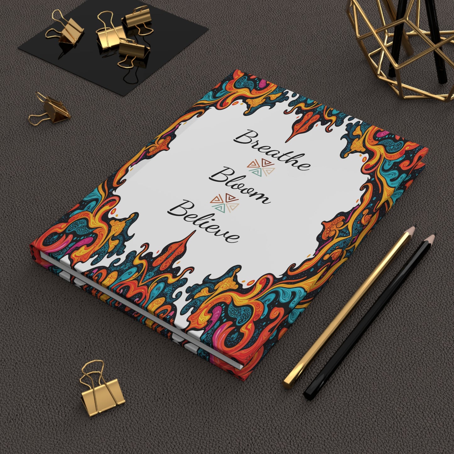 A hardcover journal with a colorful, ornate border and the words Breathe Bloom Believe on the cover sits on a textured dark surface. Gold and black binder clips, a gold geometric ornament, and two pencils (one gold, one black) surround this African-inspired notebook.