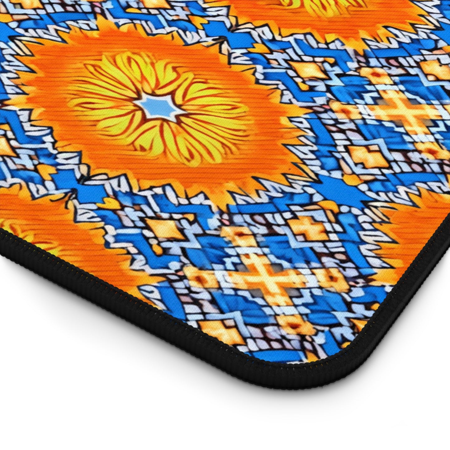 A close-up of a colorful workspace accessory, this neoprene desk mat features bright orange, yellow, and blue geometric designs. Large orange floral shapes with yellow centers are bordered by intricate blue and white patterns. The edge is black and slightly rounded.