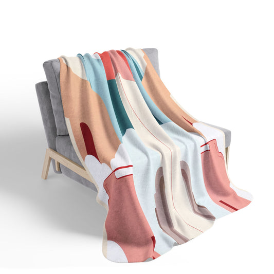 A soft, plush blanket with an abstract, African-inspired multicolored design in shades of pink, coral, beige, blue, and cream is draped over a modern light-gray upholstered bench with light wooden legs, set against a white background.