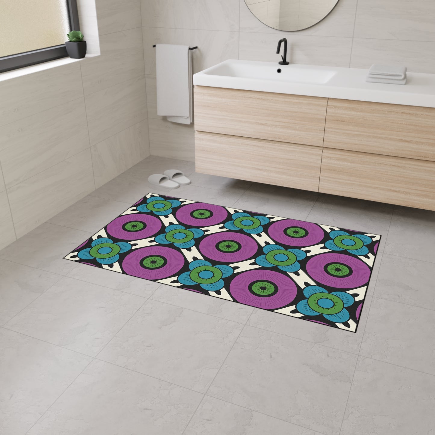 A modern bathroom with white walls and light gray floor tiles features a large, non-slip custom floor mat displaying a bold, colorful geometric flower pattern inspired by African-inspired designs beside a wooden vanity and round mirror. A towel hangs on the wall.
