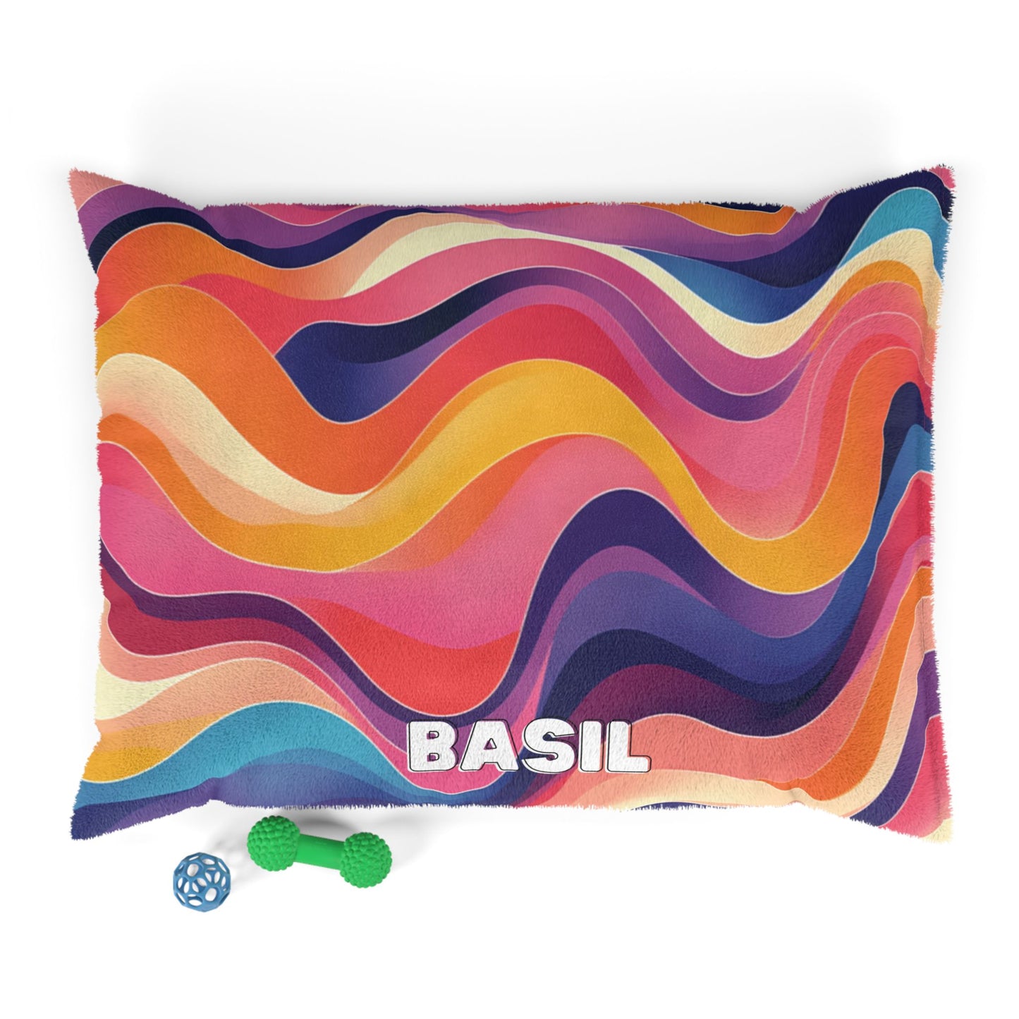 A colorful, African-inspired pet bed with wavy stripes in pink, orange, yellow, purple, and blue features the name “BASIL” in bold white letters. Two pet toys—a green bone and a blue ball—are placed below on a white background.