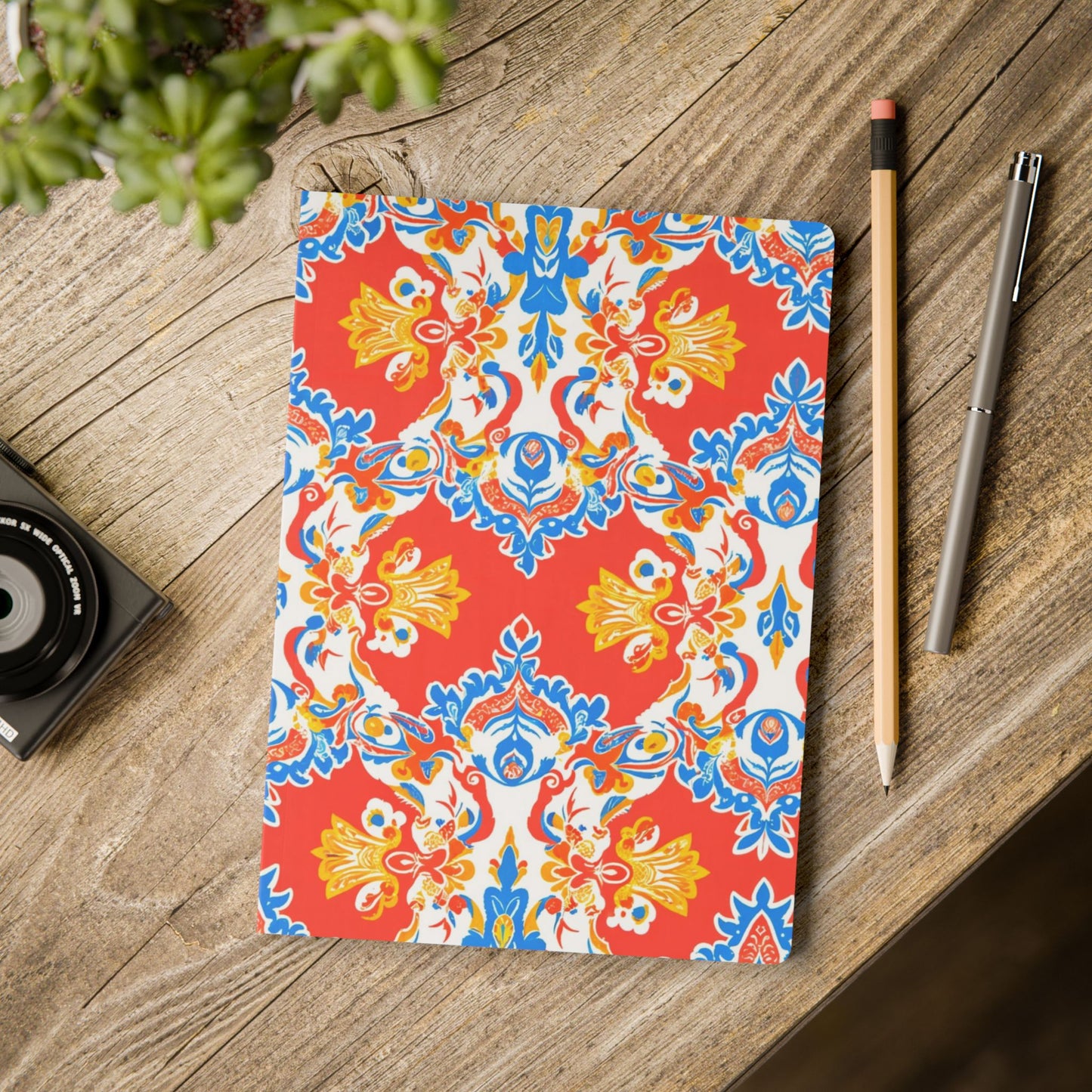 A vibrantly patterned African-inspired journal with a red, blue, yellow, and white symmetrical floral design lies on a rustic wooden table. Next to it are a pencil, silver pen, small black camera, and a potted green plant in the corner.