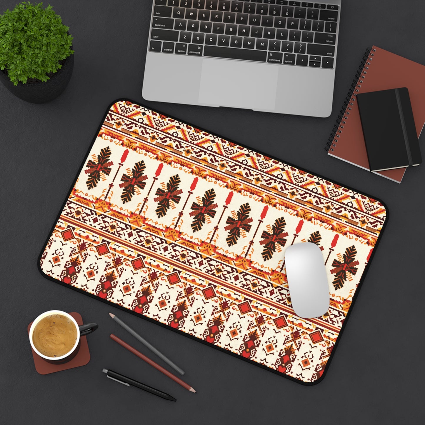 A patterned neoprene desk mat with red, brown, and black tribal-style designs sits on a dark desk beside a silver laptop, coffee cup, notebooks, plant, white mouse, and pens—a colorful workspace accessory viewed from above.