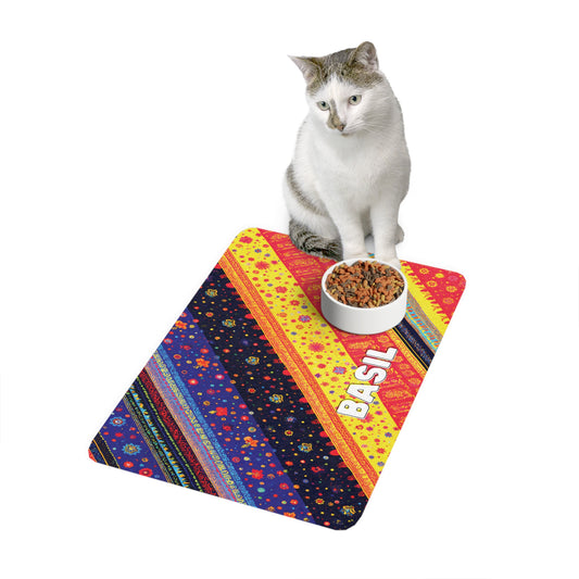 A white and gray cat sits beside a bowl of kibble on an African-inspired mat with diagonal stripes, floral patterns, and the name BASIL printed in bold white letters. The background is plain white.