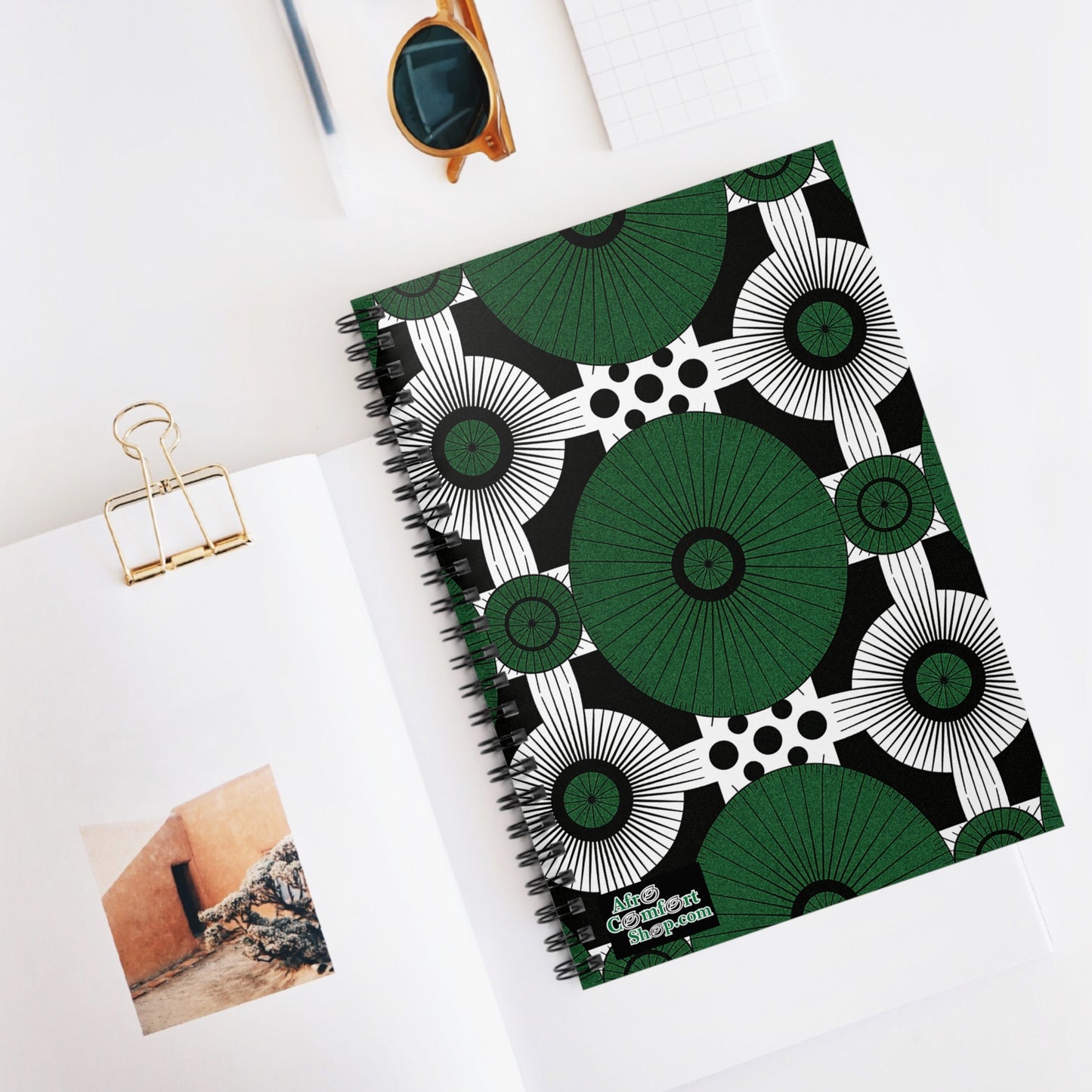 A bold green, black, and white African-inspired spiral notebook with geometric patterns lies on a white desk. Nearby are an open book with a wall photo, a gold binder clip, tortoiseshell sunglasses, and a white notepad.