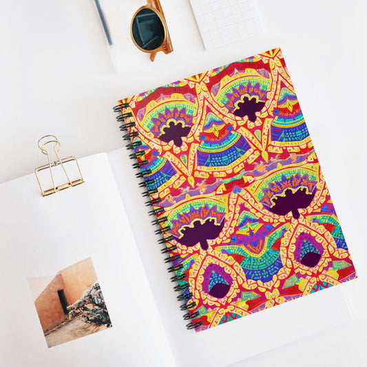 A colorful African-inspired spiral notebook with a vibrant, symmetrical geometric pattern in red, yellow, green, and blue lies on a white desk. Nearby are an open photo book, a gold binder clip, sunglasses, and a small grid notepad.