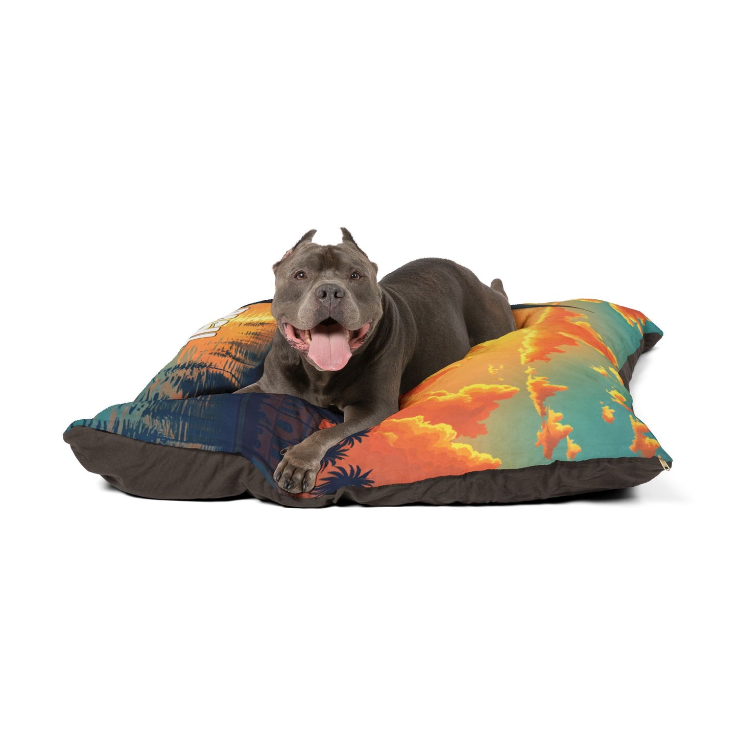 A gray pitbull with cropped ears and a pink tongue out lies happily on a colorful pillow featuring tropical sunset and palm tree designs, showcasing luxurious comfort against a white background.