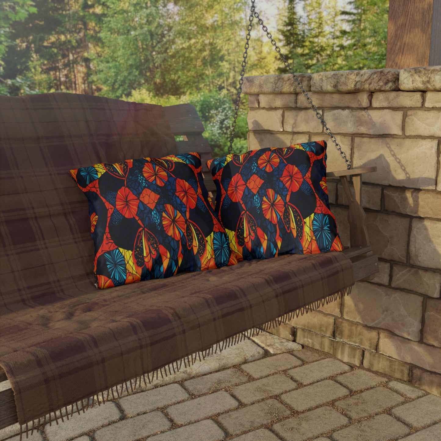 A wooden porch swing with a plaid blanket and two UV-resistant outdoor pillows featuring red, orange, blue, and yellow leaf patterns sits beside a stone wall. The brick patio and trees outside are visible in the background.
