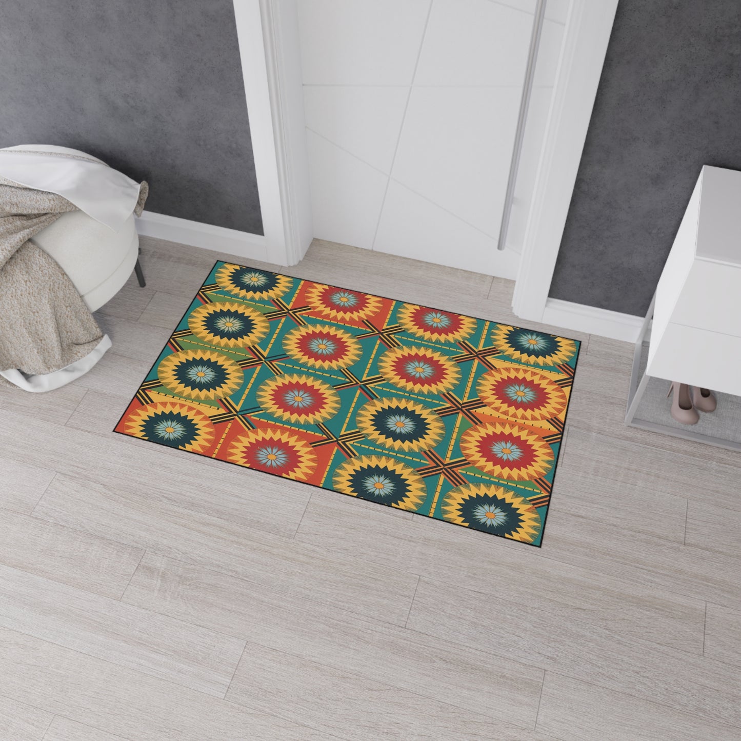 A colorful African-inspired floor mat with a geometric starburst pattern in red, yellow, green, and blue sits on light wooden flooring by a white doorway. It features non-slip rubber backing to keep it securely in place.