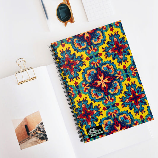 A colorful African-inspired spiral notebook with an ornate, floral, red and blue tile pattern on a yellow background rests on a white desk beside an open book with a photo, gold binder clip, grid notepad, and brown sunglasses.