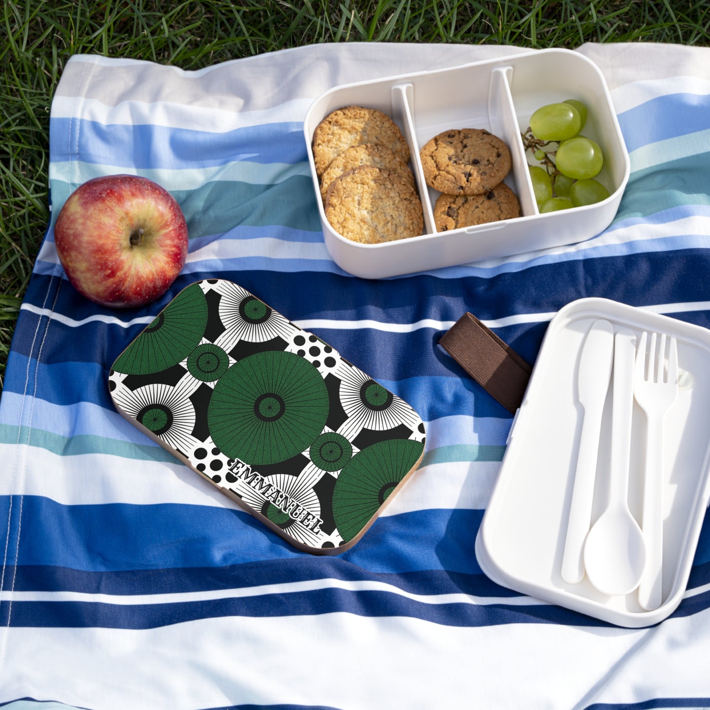 A striped blue and white picnic blanket on grass holds an apple, a BPA-free lunchbox with cookies and grapes, and another open lunchbox with utensils. A green patterned personalized lunch box labeled “EMMANUELLE” sits nearby.