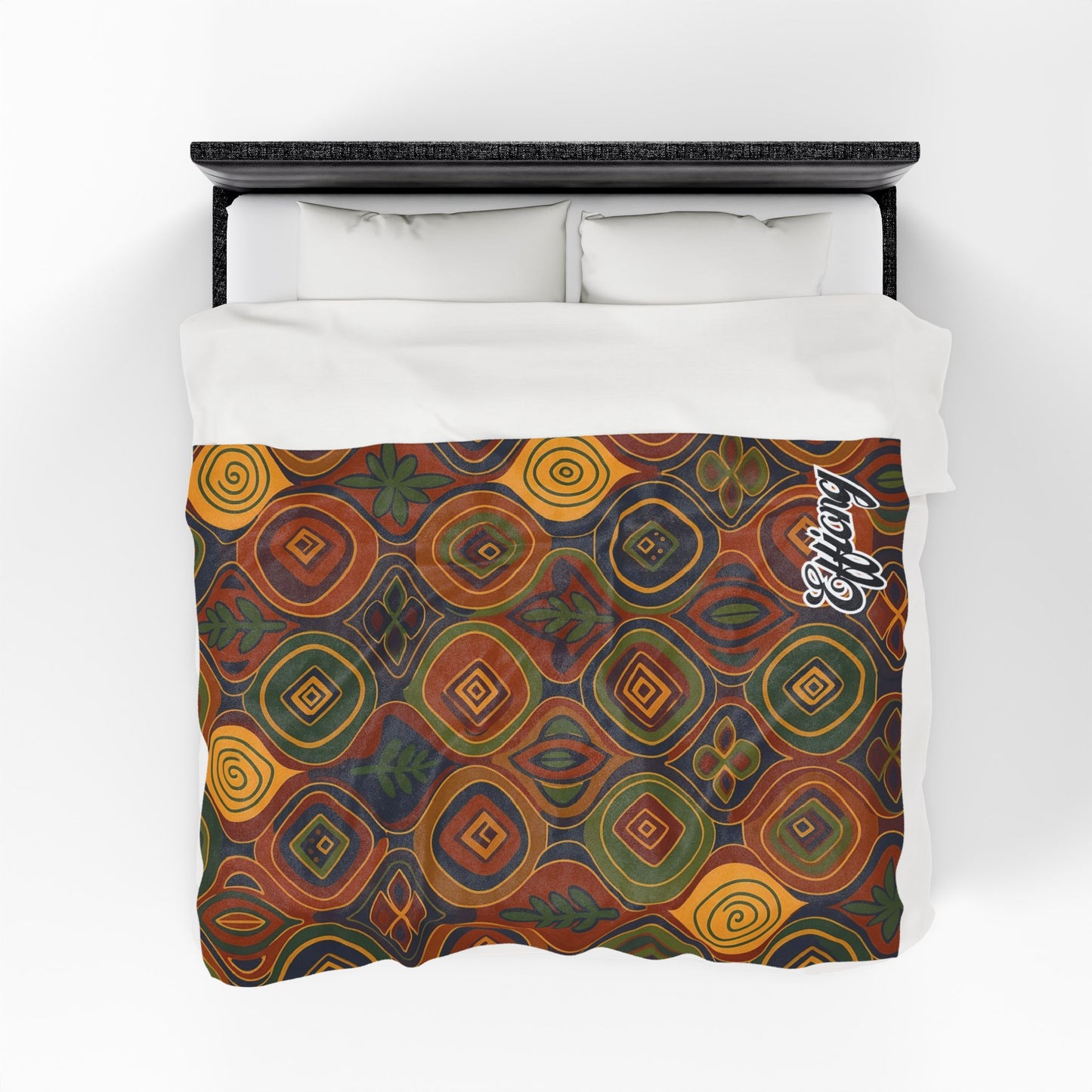 A bed with white pillows and a white sheet is covered by an African-inspired blanket featuring concentric diamond and circle patterns in earthy tones, leaves, flowers, and the word Legacy scripted in white in the corner.