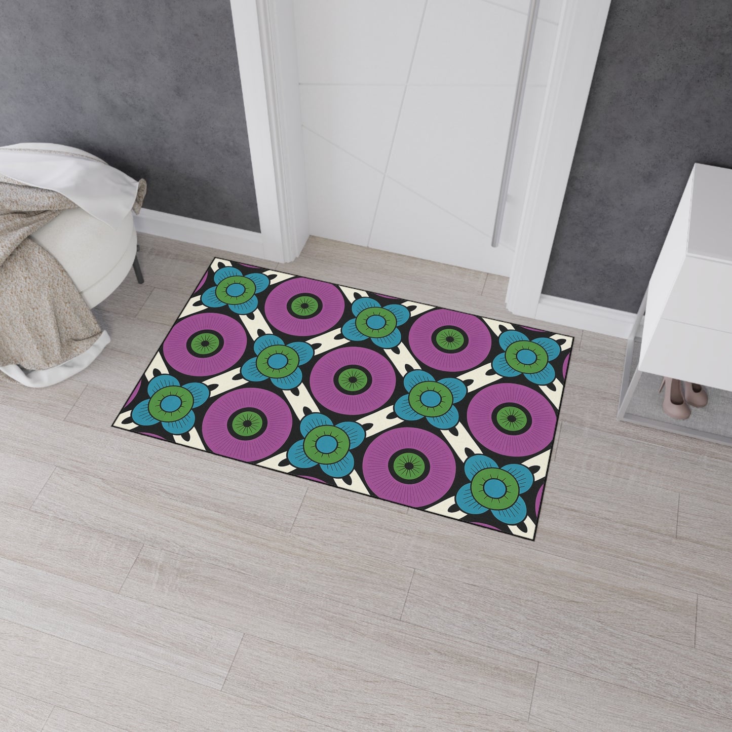 Rectangular rug on light wood floor features a bold, African-inspired geometric pattern with concentric circles in purple, turquoise, and green, separated by black lines on a cream background. The rug is near a white cabinet and beige throw blanket.