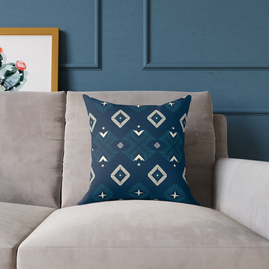 A square, African-inspired pillow with a blue background and white geometric tribal patterns rests on a light gray sofa. The wall behind is blue with molding, and a framed cactus print with a wooden frame is partially visible on the left.