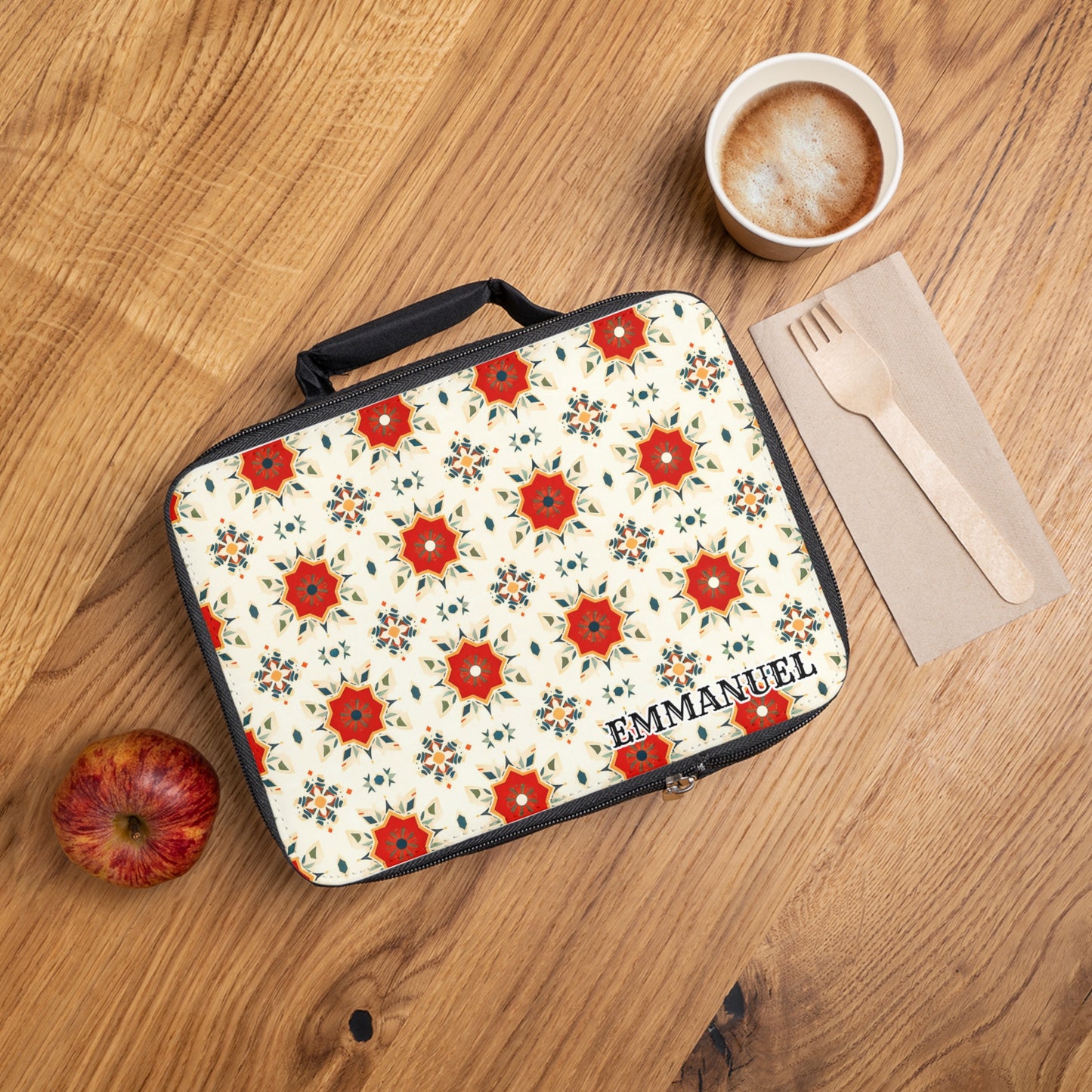 A personalized lunch bag labeled “EMMANUEL” sits on a wooden table with a red apple, latte with foam, and utensils. The insulated lunch bag features a red and floral star design on a cream background.