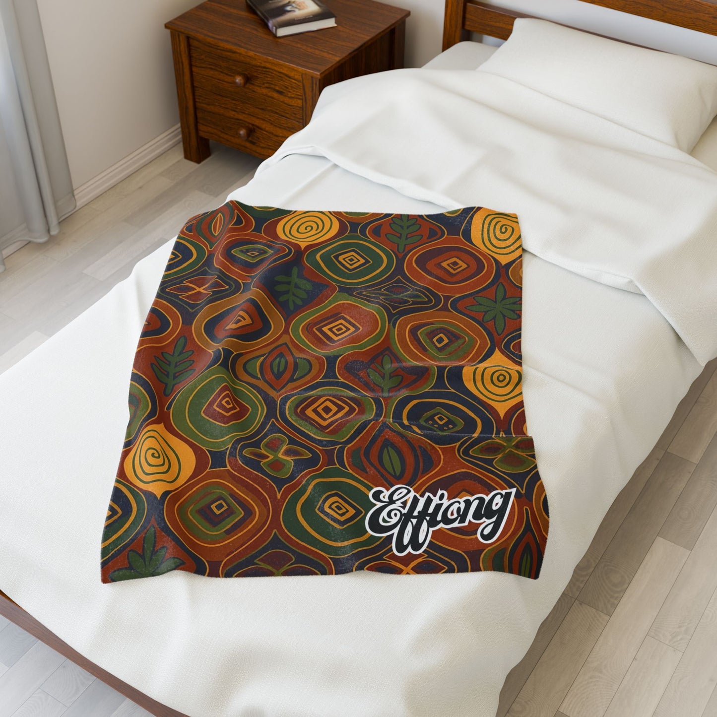 A colorful, African-inspired blanket with wavy geometric patterns in orange, green, yellow, and brown is spread over a white-covered bed. This personalized velveteen blanket has “Effong” written in cursive on one corner by the wooden nightstand.