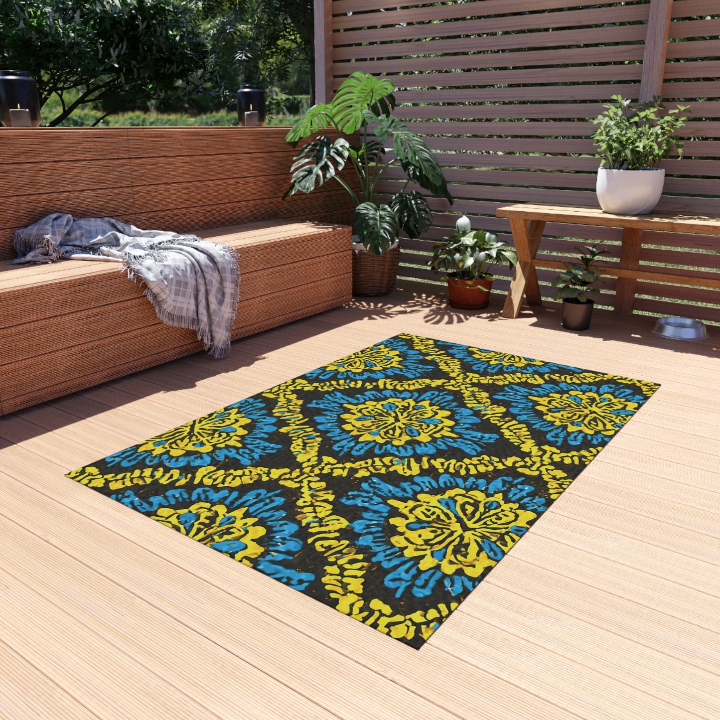 A cozy outdoor patio with light wooden decking features a wicker sofa topped with gray cushions and a plaid blanket. An African-inspired outdoor rug in blue and yellow patterns brightens the floor, while potted plants and a slat screen complete the relaxing scene.