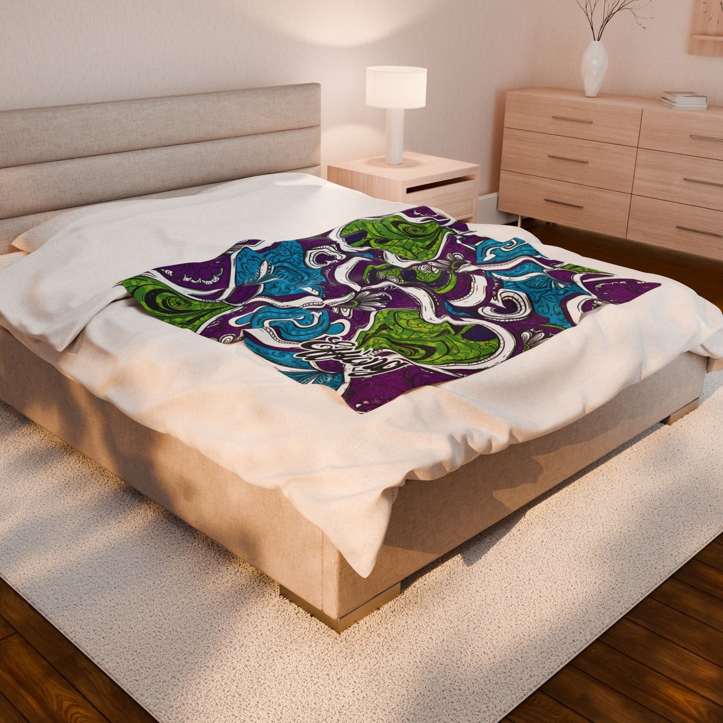 A modern bedroom with a beige bed and light wood furniture. A bright, African-inspired blanket in green, blue, purple, and white is spread over the bed, contrasting with the plain white bedding and neutral decor.