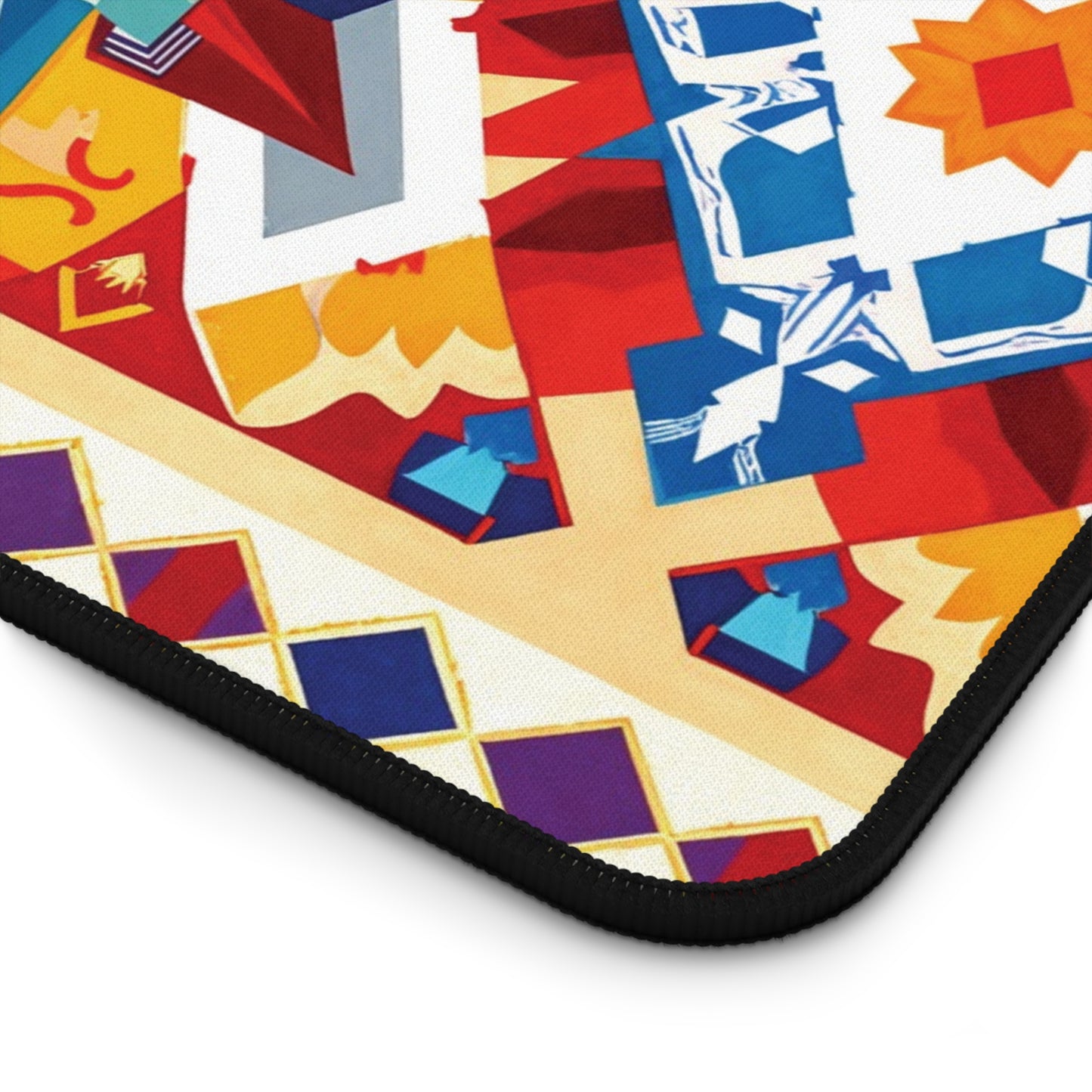 A close-up of a colorful, geometric-patterned neoprene desk mat with red, blue, yellow, white, and purple shapes—squares, diamonds, and stylized floral designs. The black fabric edge of this vibrant workspace accessory is visible at the lower left.