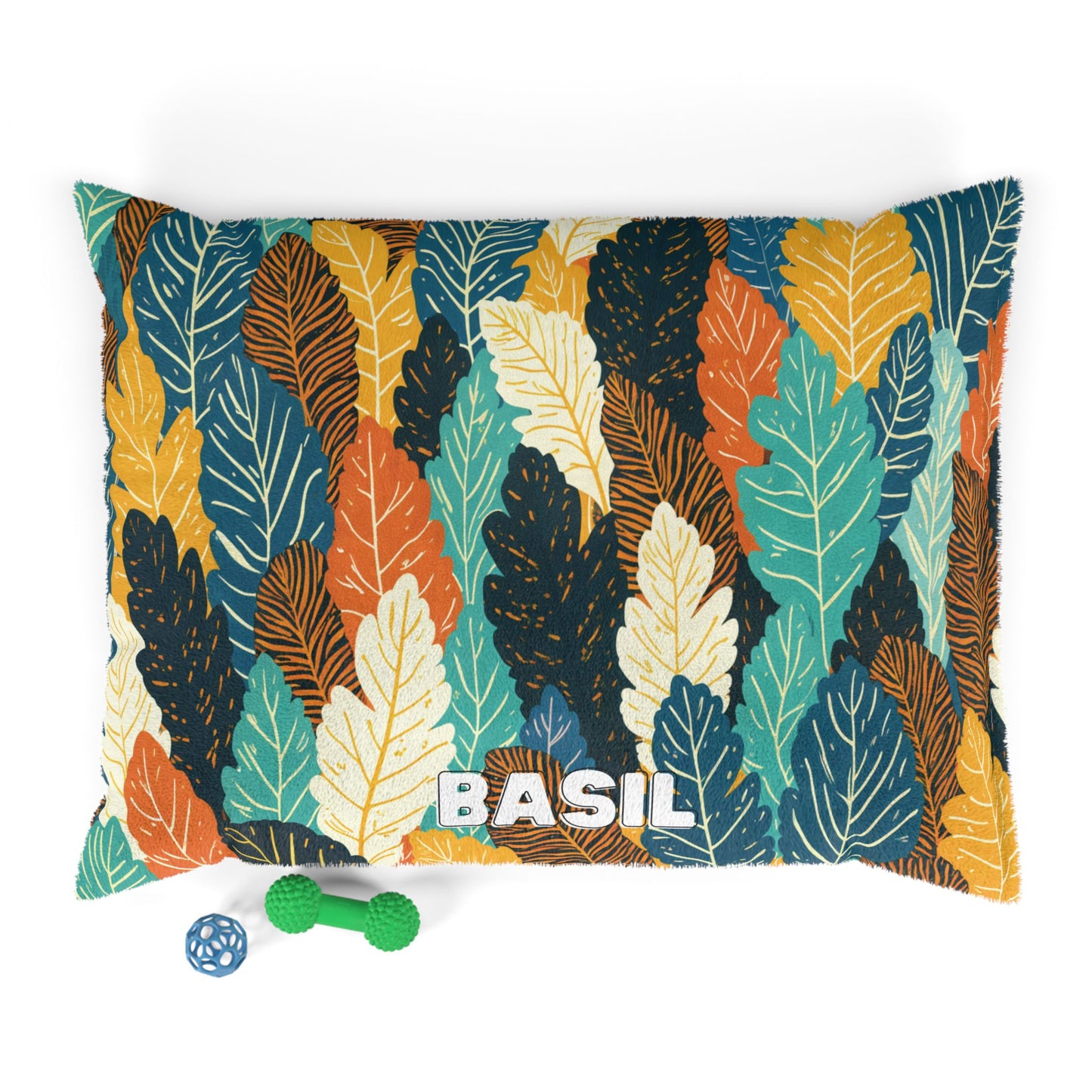 A rectangular personalized pet bed with a colorful leaf-patterned cover in teal, yellow, orange, cream, and black. The name BASIL is printed in bold white letters. Two pet toys rest nearby on a white background. Durable construction ensures lasting comfort.