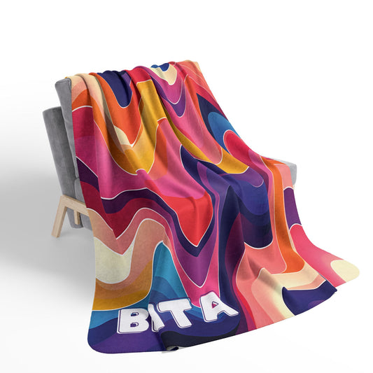 A gray armchair is draped with a colorful sherpa fleece blanket featuring wavy, abstract patterns in red, orange, blue, yellow, and pink. The lower corner displays bold white letters partially spelling “BITA.” The minimal setting is well-lit.