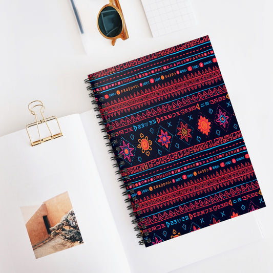 A premium writing notebook with a colorful geometric and floral pattern on a dark cover lies on a white desk. Nearby are an open photo book, a gold binder clip, sunglasses, and a small notepad, creating a stylish, organized workspace scene.