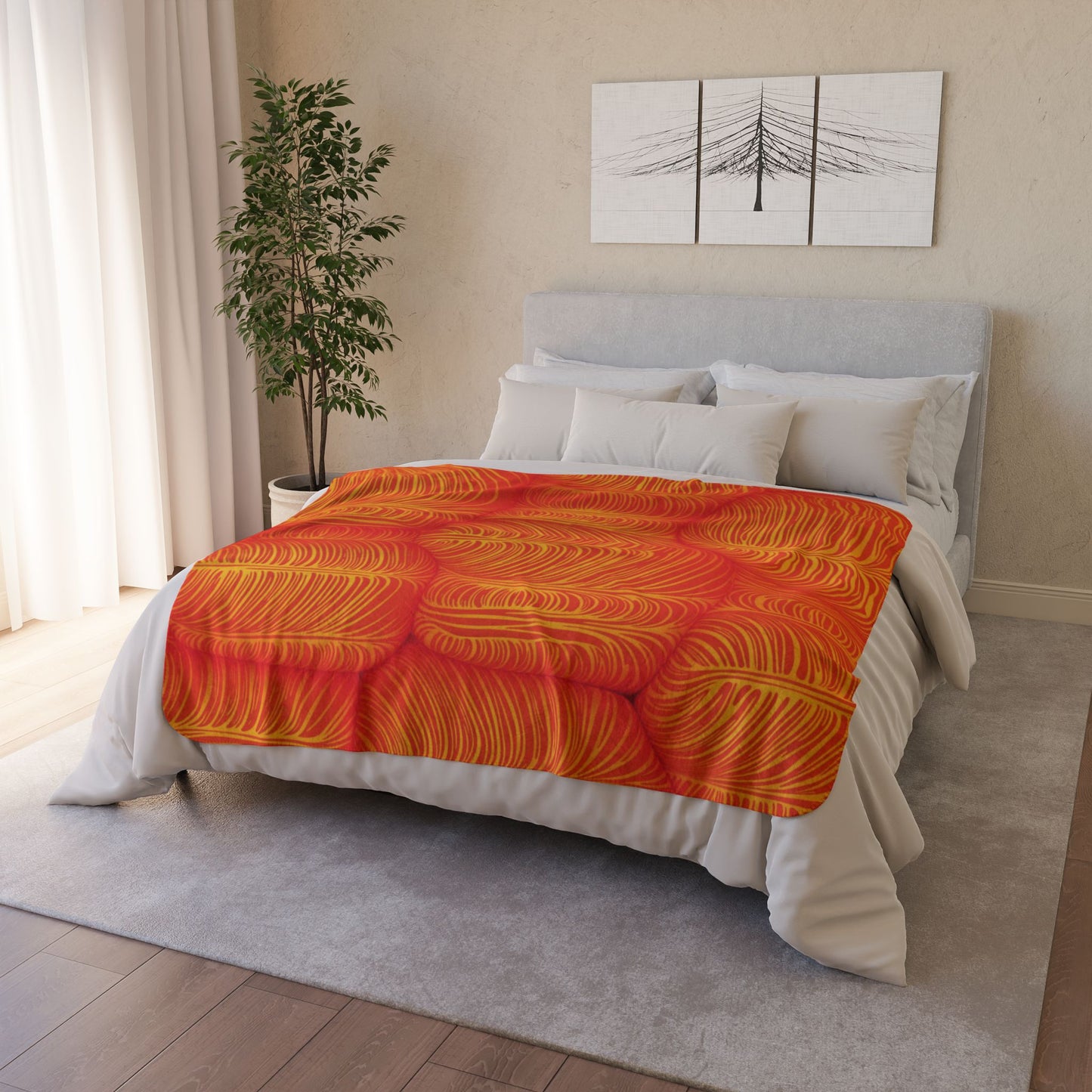 A neatly made bed with a light gray headboard is covered by an African-inspired fleece blanket in vibrant orange and yellow patterns. White pillows accent the blanket, while cozy home decor touches like a green plant complete the space.