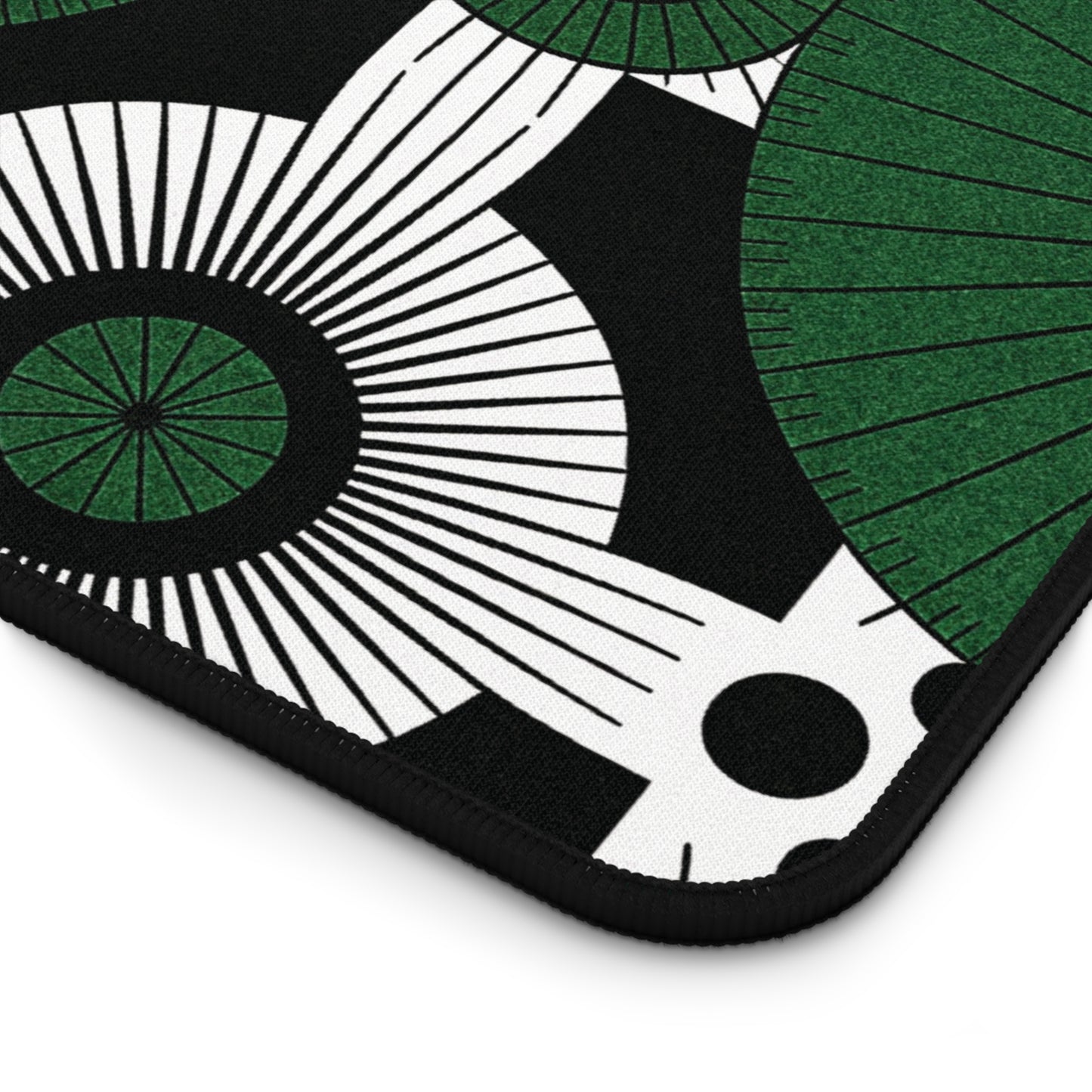 A close-up of an ergonomic desk pad featuring a modern, abstract pattern. Green and white circular shapes with radiating lines, black centers, and black polka dots stand out on the black background. The neoprene desk mat has rounded, black-stitched edges.