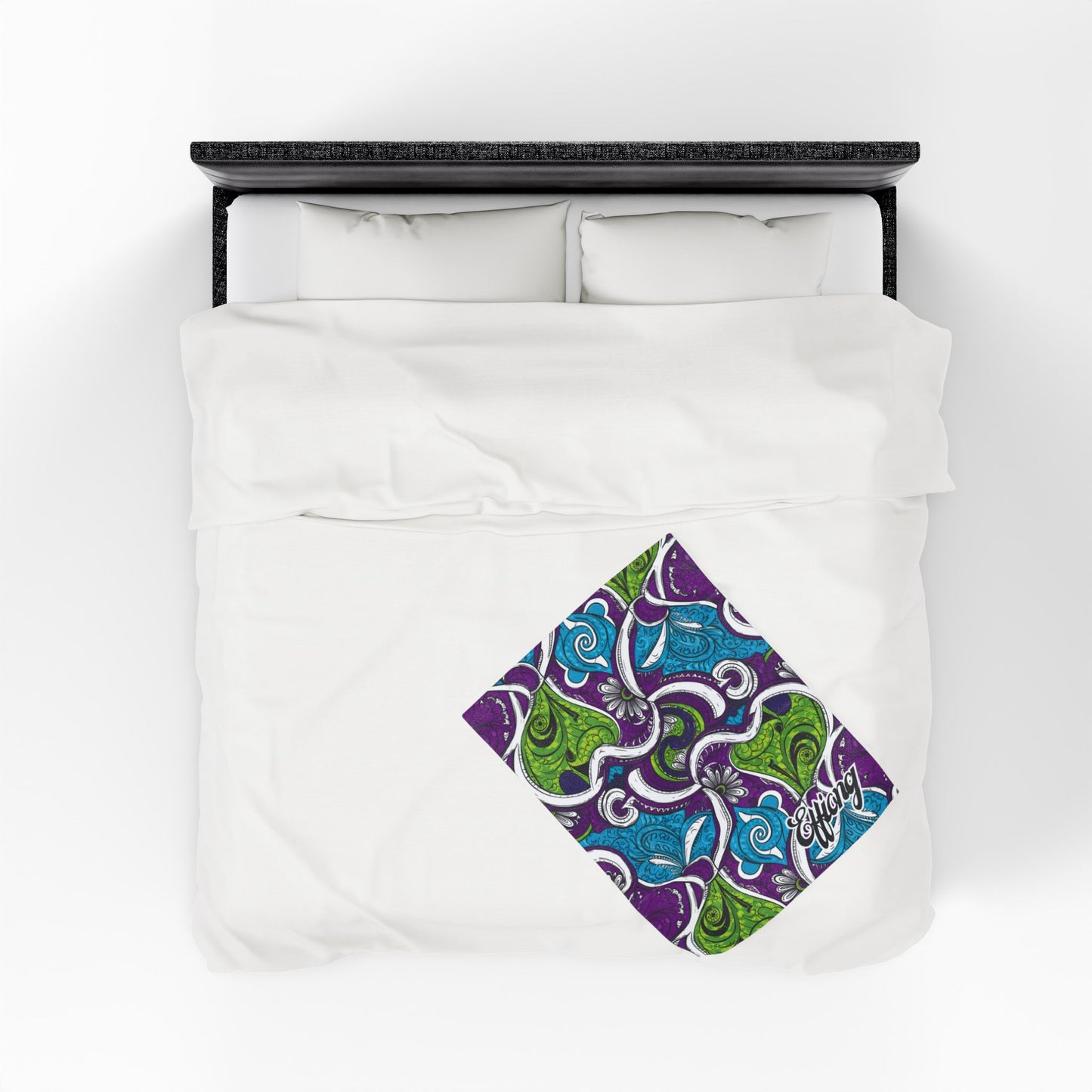 A neatly made bed with white bedding is shown from above. An African-inspired blanket with a colorful, square-patterned design in blue, green, and purple swirls decorates the bottom right corner of the comforter. Two pillows rest at the head of the bed.