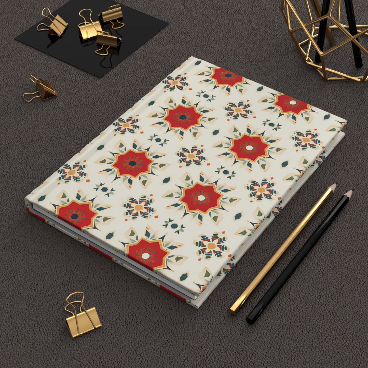A closed African-inspired hardcover journal with a white cover and an ornate red, orange, and green geometric floral pattern rests on a textured dark surface. Nearby are gold binder clips, a gold decor piece, and pencils.