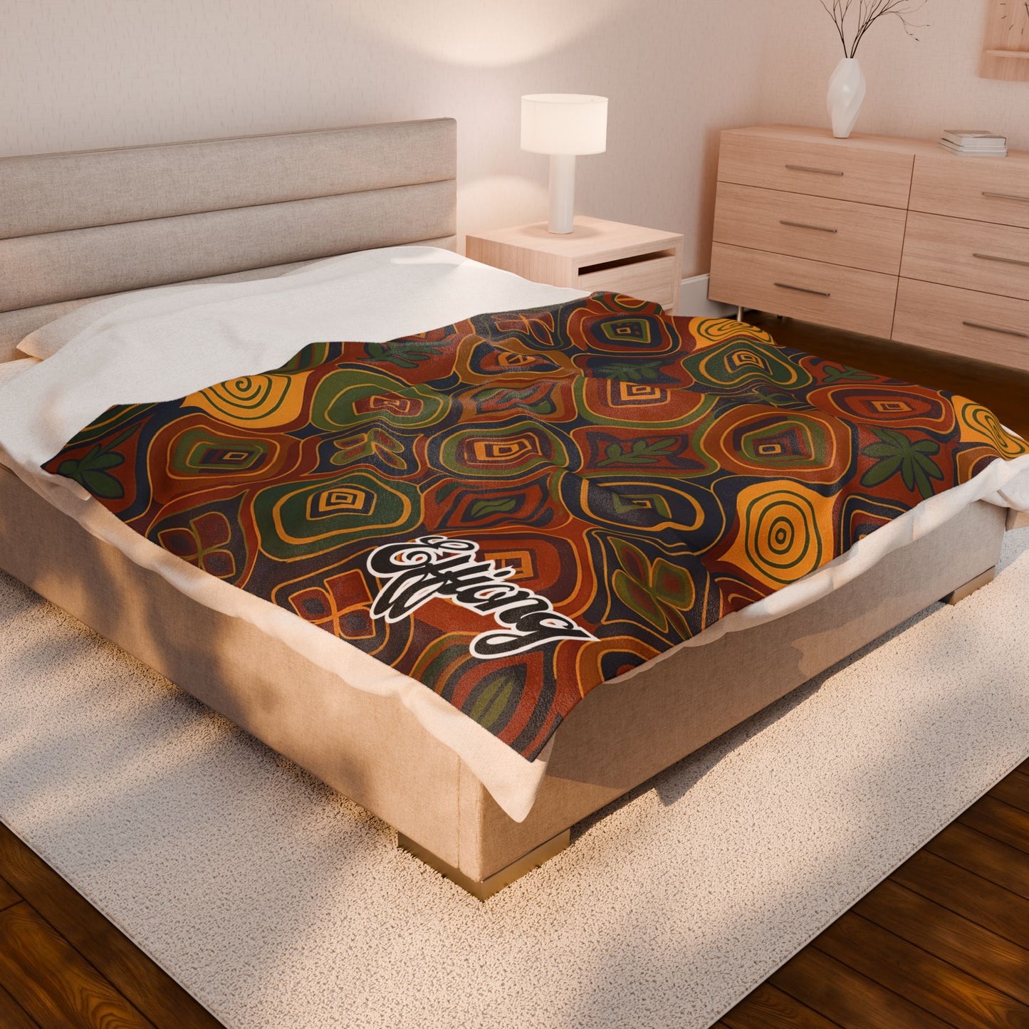 A neatly made bed with a colorful, African-inspired blanket in warm shades of red, orange, yellow, and green, featuring a white script logo in one corner. The room has a beige headboard, wooden furniture, white lamp, and light décor.