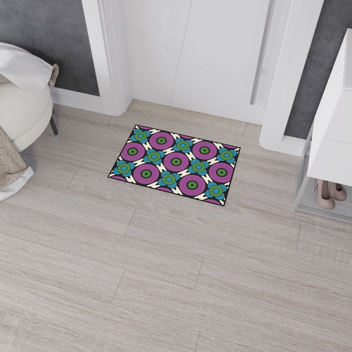 A rectangular doormat with a colorful geometric pattern and African-inspired aesthetics features purple circles, blue flowers, and green centers. Its non-slip rubber backing keeps it secure on light wood flooring in a modern entryway near a white cabinet and bed.