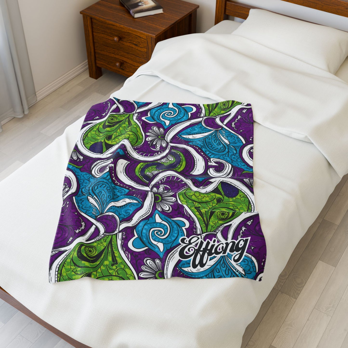 A single bed with white bedding and a wooden nightstand beside it. A personalized velveteen blanket featuring green, blue, and purple abstract paisley patterns and the word Effing is spread across the lower half of the bed.