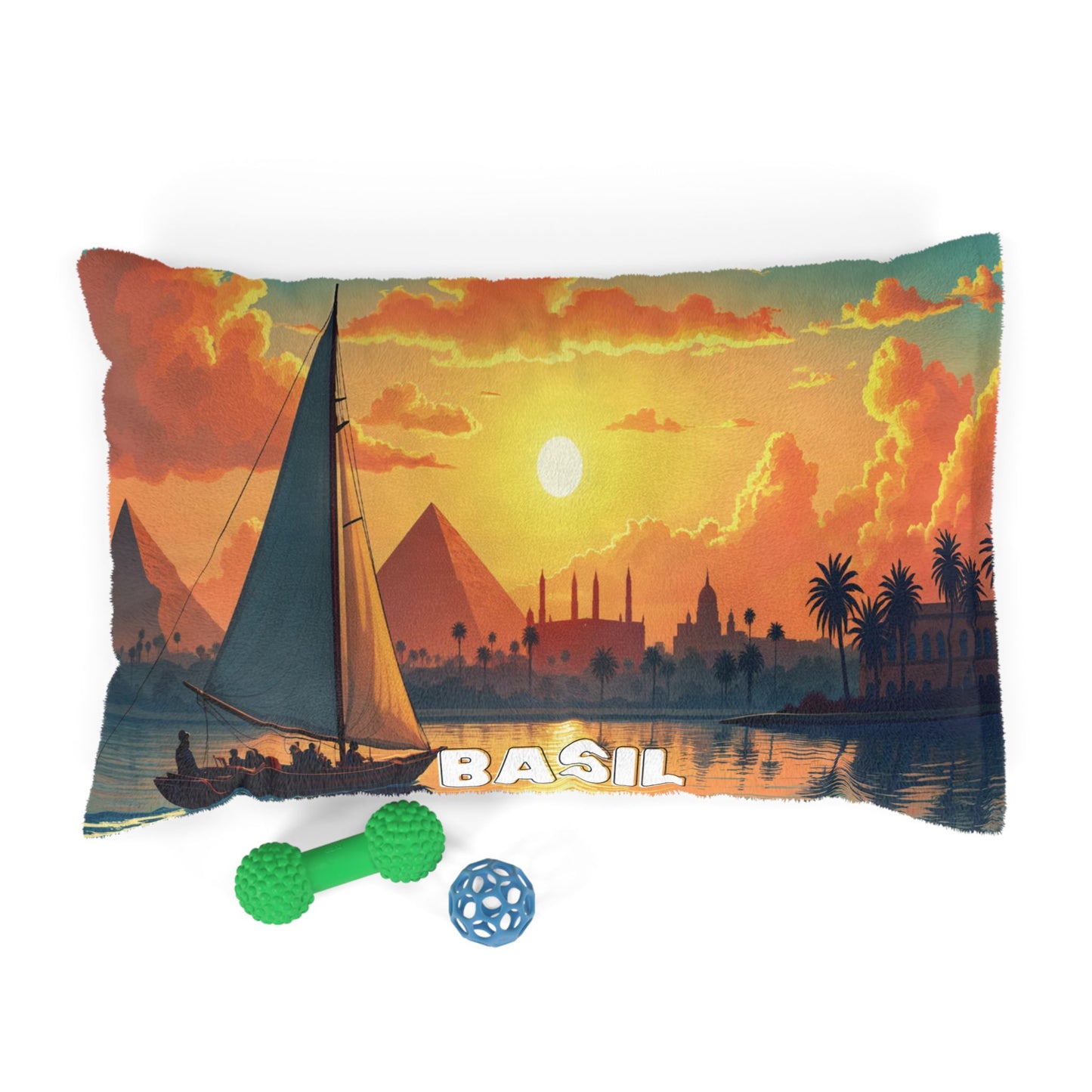 A rectangular pillow with a vivid sunset scene of pyramids, palm trees, and a sailboat on water; “BASIL” is written in bold white letters. In front are a green dumbbell-shaped dog toy and a blue ball toy—perfect for personalized pet beds.