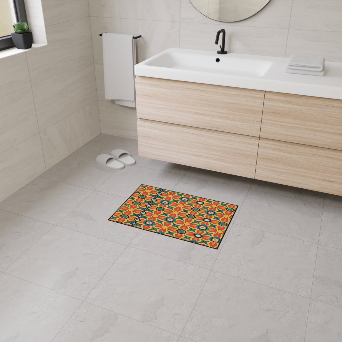 A bright, multicolored mat with a geometric floral pattern and non-slip rubber backing is placed on a light gray tiled bathroom floor. Nearby are white slippers, a modern wood vanity with a sink, a round mirror, and a white hand towel hanging by the wall.