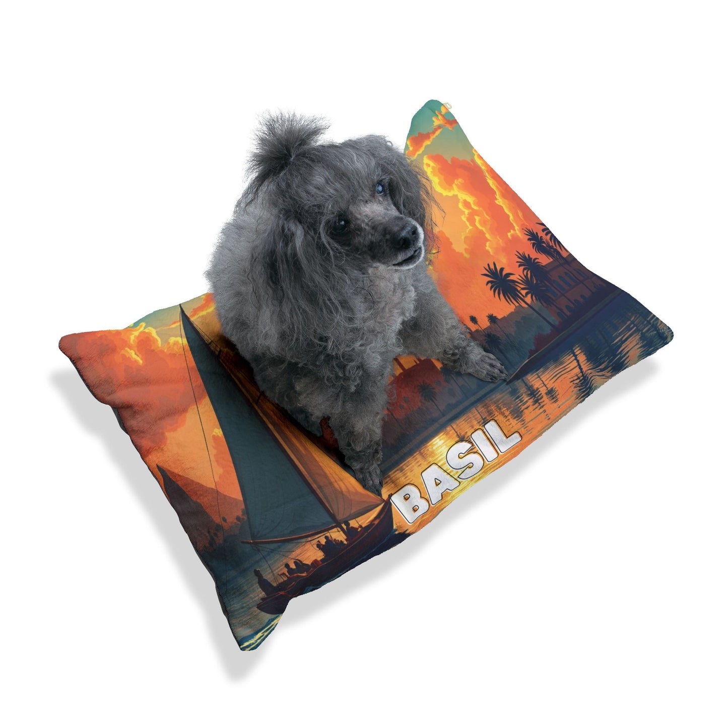 A small gray poodle with curly fur sits on a personalized pet bed—a pillow printed with an orange sunset, palm trees, a sailboat, and BASIL in bold letters. The plain white background highlights the luxurious comfort of this unique pet accessory.