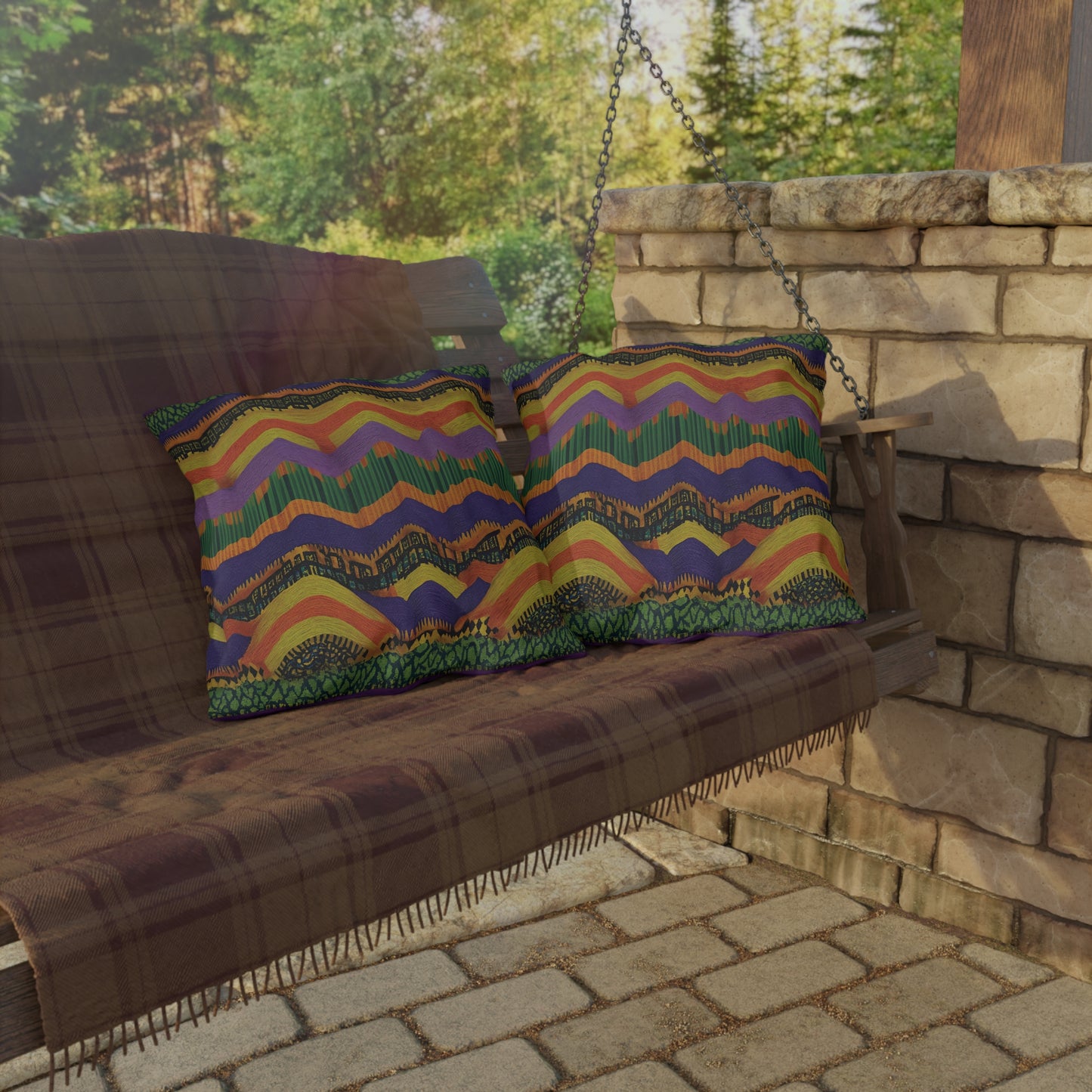 A wooden porch swing covered with a plaid blanket holds two colorful outdoor pillows made from UV-resistant fabric. The swing hangs by chains beside a stone wall, and sunlight filters through surrounding trees onto the brick patio floor.