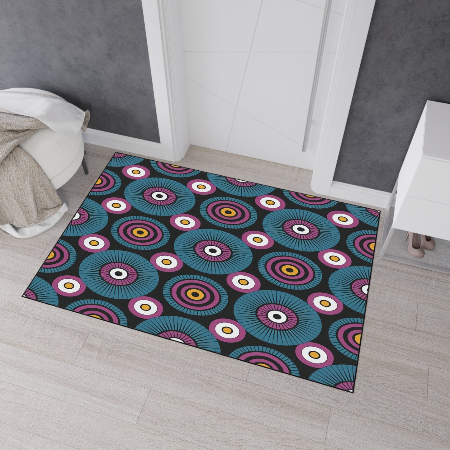A colorful, African-inspired rug with concentric circle patterns in teal, purple, pink, white, orange, and black lies on a light wood floor in front of a white door. A beige blanket is draped on the left; a white cabinet peeks in on the right.