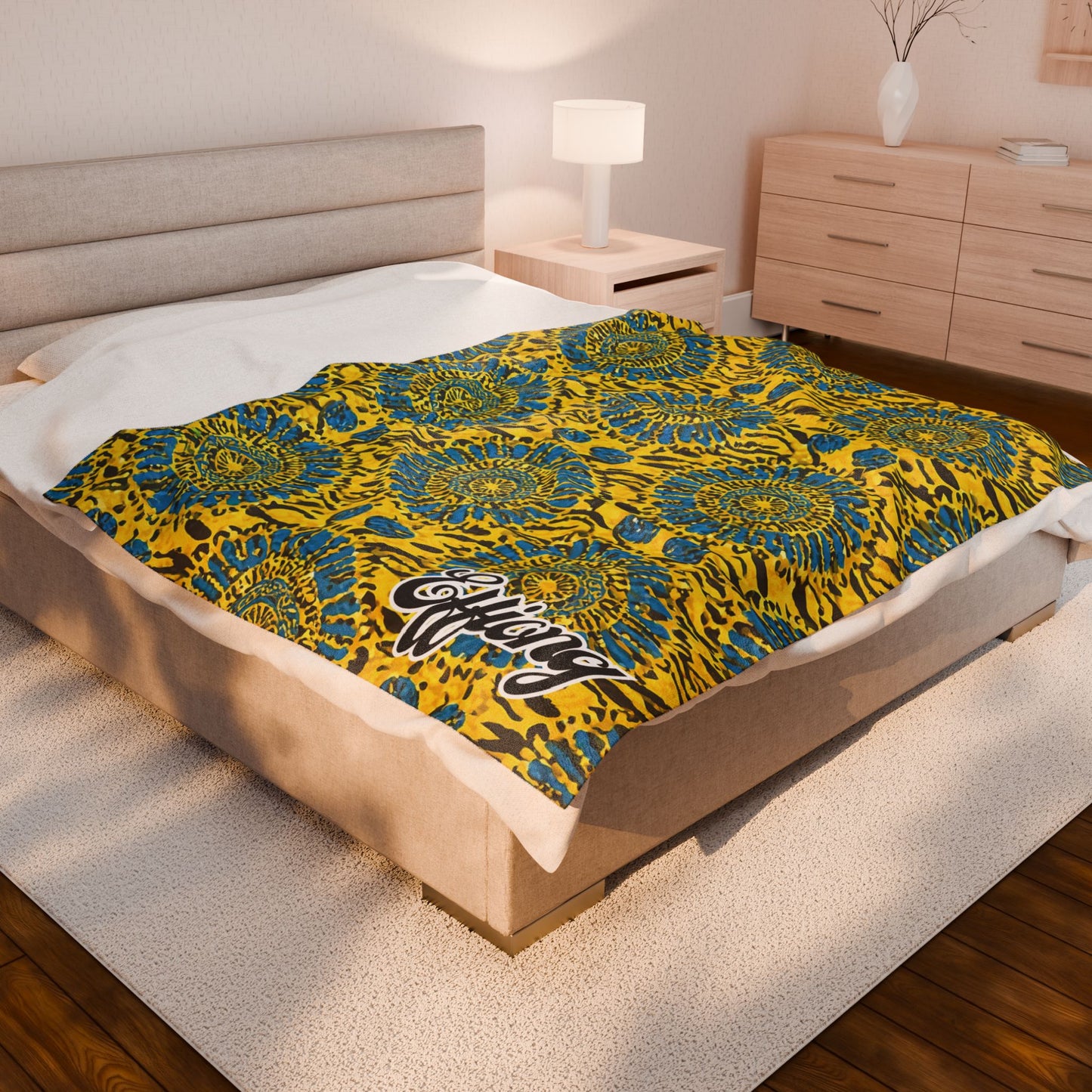 A beige bed with a personalized velveteen blanket featuring yellow, blue, and black abstract floral and animal print patterns, plus white script text in one corner. The room has light wood furniture, a lamp, and a vase with branches on a dresser.