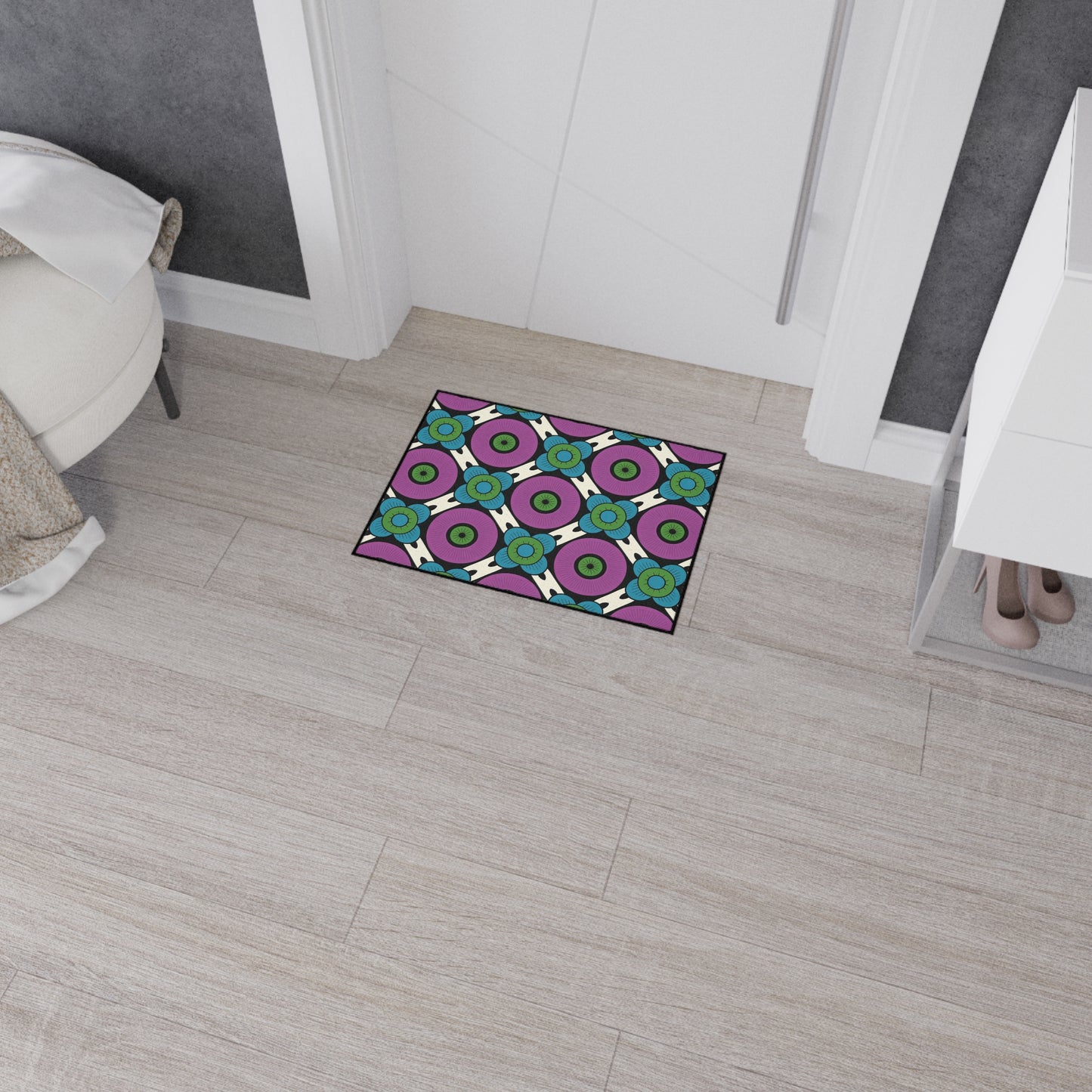 A small, rectangular mat with a colorful, African-inspired geometric pattern in purple, green, blue, black, and white lies on light wood flooring in a modern room. This custom floor mat features a non-slip rubber backing for added safety.