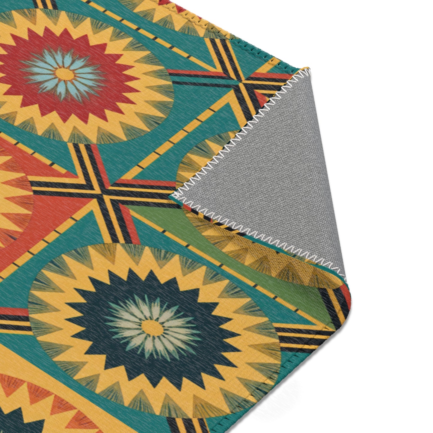 A close-up of a colorful African design rug with bold geometric and starburst patterns in teal, red, yellow, and black. A corner is flipped over, revealing the plain gray underside and zigzag white stitching along the edge.