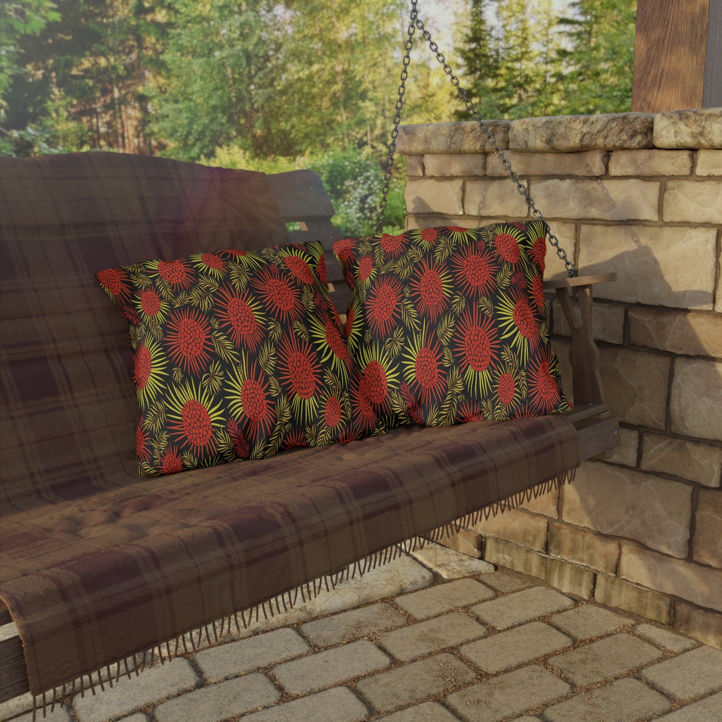 A wooden porch swing with a plaid blanket and two African-inspired square cushions featuring a red and yellow floral pattern sits next to a stone column. Sunlight filters through the trees onto the brick patio, creating a cozy outdoor setting.
