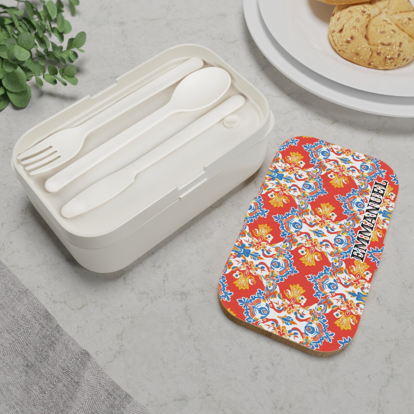 A personalized African-inspired bento lunch box with a fork, spoon, and knife sits on a light stone surface. Its colorful lid, boldly labeled “EMMANUEL,” rests beside plates of cookies and a lush leafy plant.